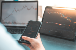 a hand hold a phone with stocks with a laptop and tablet looking at stocks